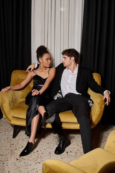 Young african american woman in black dress and handsome man in elegant suit sitting on velvet sofa — Stock Photo