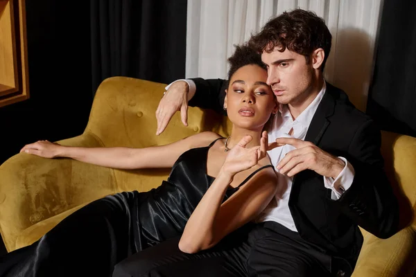 Giovane coppia multiculturale in elegante abbigliamento che si toccano le dita e si siedono sul divano di velluto — Foto stock