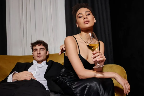 Young african american woman holding glass of wine and sitting with man in suit on velvet sofa — Stock Photo