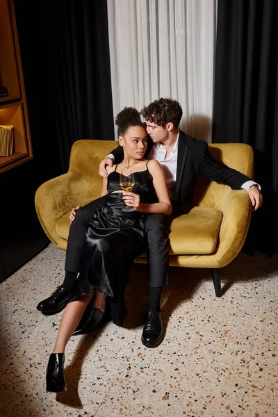 Кучерява афроамериканка тримає келих вина і фліртує з чоловіком у костюмі на оксамитовому дивані — стокове фото