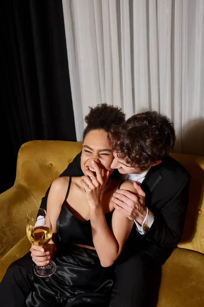 Cheerful african american woman holding glass of wine and laughing near man in suit on velvet sofa — Stock Photo