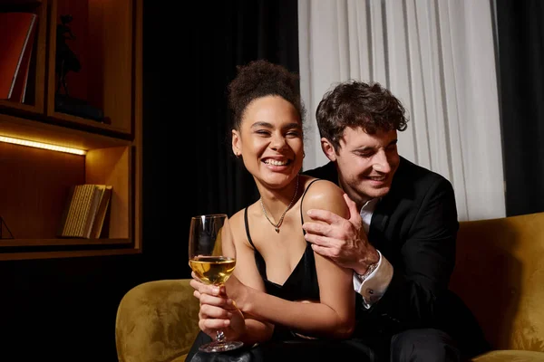 Man hugging excited african american woman holding glass of wine and sitting on velvet sofa — Stock Photo