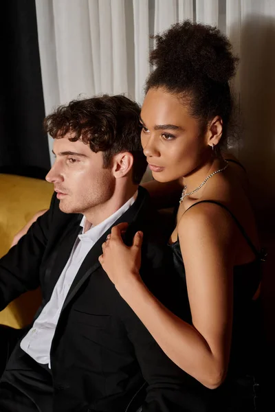 Attractive african american woman in dress hugging man in formal wear and posing on velvet sofa — Stock Photo