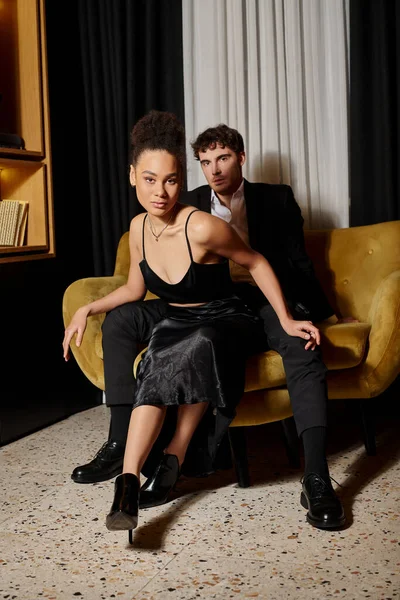 Young multicultural couple in black evening attire posing together while sitting on velvet sofa — Stock Photo
