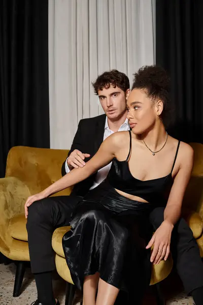 Casal multicultural em roupas pretas noite posando juntos enquanto sentado no sofá de veludo — Fotografia de Stock