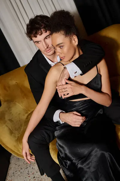 Pareja multicultural en traje de noche posando juntos mientras están sentados en un sofá de terciopelo, elegancia - foto de stock