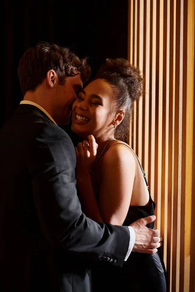 Momento íntimo de pareja multicultural en negro noche desgaste abrazo y sonriendo durante la fecha - foto de stock