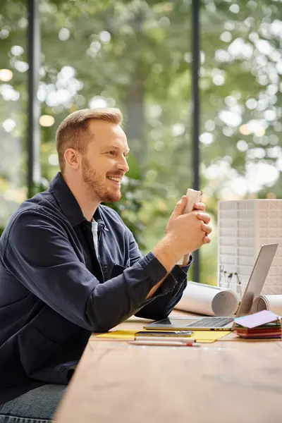 Cheerful handsome man in everyday attire sitting at desk and working on his startup while in office — Stock Photo