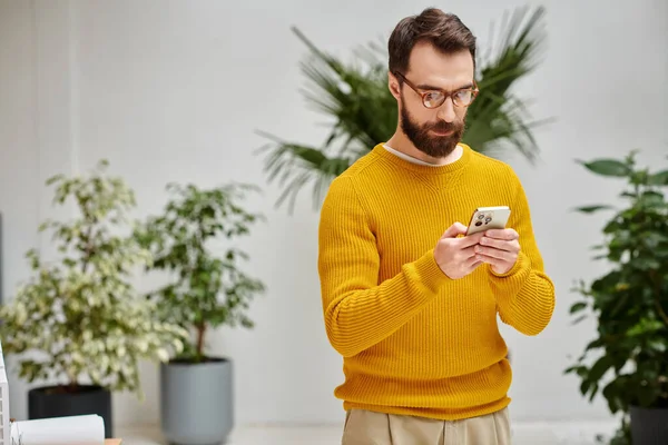 Hombre barbudo atractivo concentrado en cuello alto amarillo sosteniendo su teléfono móvil en la oficina - foto de stock