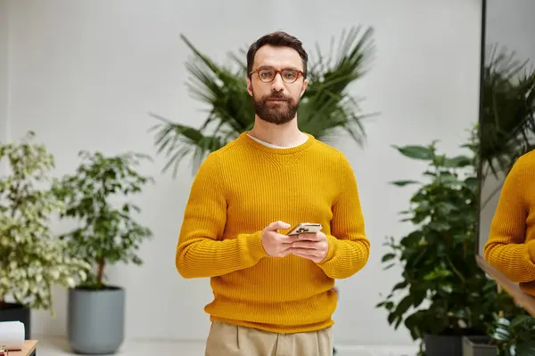 Concentrado atraente barbudo homem no amarelo gola alta segurando telefone celular e olhando para a câmera — Fotografia de Stock