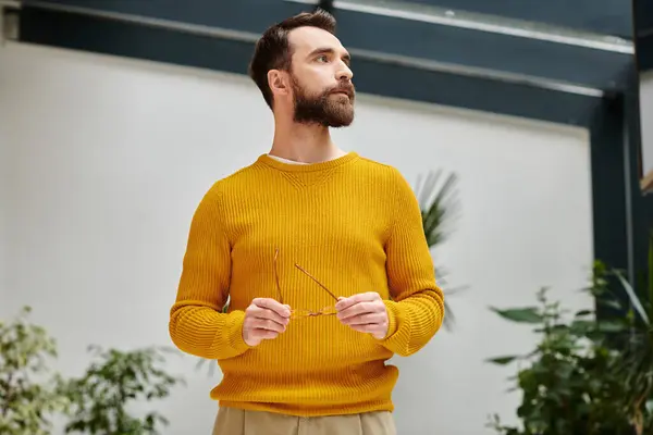 Concentrated attractive bearded man in yellow turtleneck holding his glasses and looking away — Stock Photo