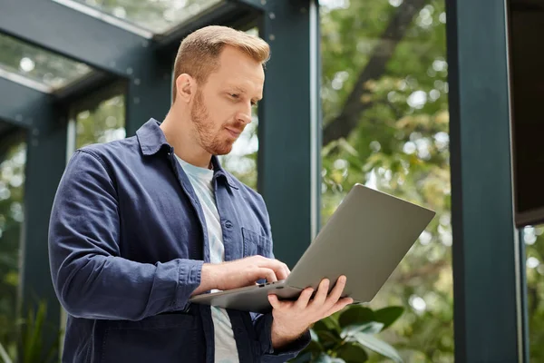 Focused handsome man in everyday comfy attire holding his laptop while working hard in office — Stock Photo