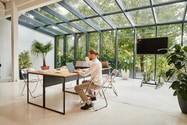 Handsome young man with beard in white casual turtleneck sitting in his office and working on laptop — Stock Photo