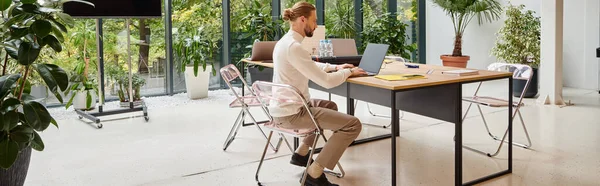 Bell'uomo con la barba in dolcevita bianco seduto nel suo ufficio e che lavora su laptop, banner — Foto stock