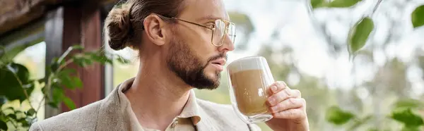 Красивий зосереджений чоловік з бородою і зібраним волоссям в елегантному костюмі, що п'є свою гарячу каву, банер — стокове фото