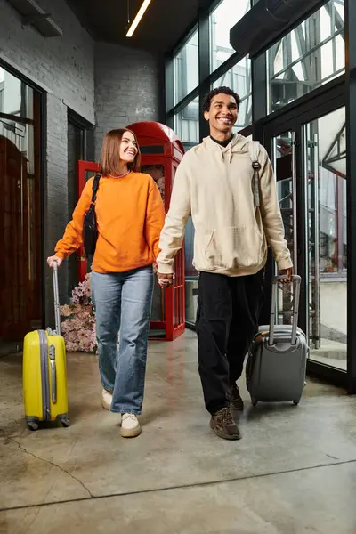 Interracial feliz pareja camina a través del hall de entrada del albergue, mientras que tirar de equipaje y tomarse de la mano - foto de stock