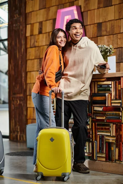 Різноманітна усміхнена пара стоїть поруч з їхньою жовтою валізою, одягнена в стильний одяг біля книг — стокове фото