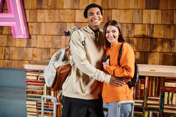 Feliz hombre y mujer diversos rodeados de libros, de pie cerca del equipaje y listo para el próximo viaje - foto de stock