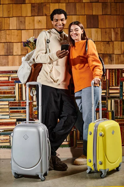 Різноманітна щаслива пара, що ділиться моментом радості, коли вони дивляться на смартфон у хостелі, готові до поїздки — стокове фото