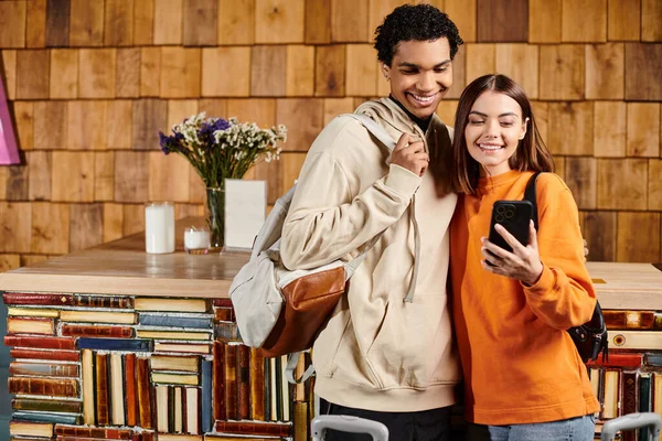Різноманітна щаслива пара, що ділиться моментом радості, коли вони дивляться на смартфон у хостелі, подорожують — стокове фото