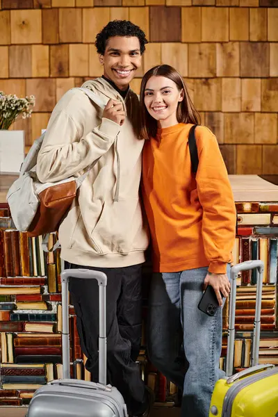 Interracial feliz pareja de pie con teléfono inteligente y equipaje cerca de estantes de libros en el albergue, viajes - foto de stock