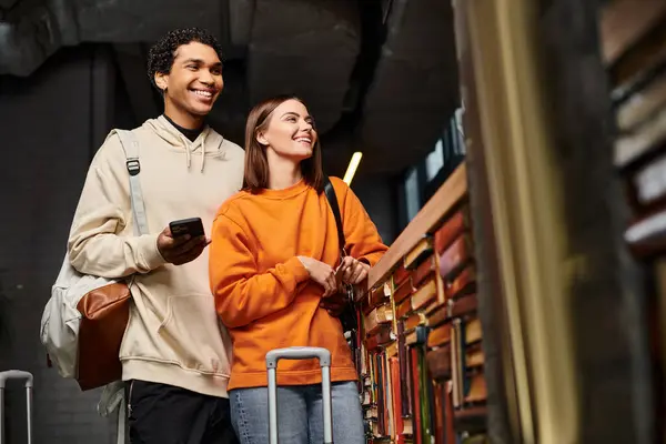 Щаслива і різноманітна пара зі смартфоном, що розділяє радісний момент у хостелі біля книжкової полиці — стокове фото