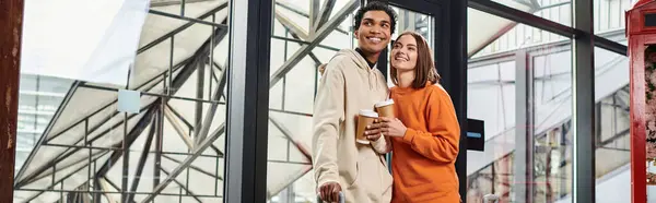 Joven pareja diversa sonriendo mientras sostiene el café para ir y entrar en un albergue moderno, pancarta - foto de stock