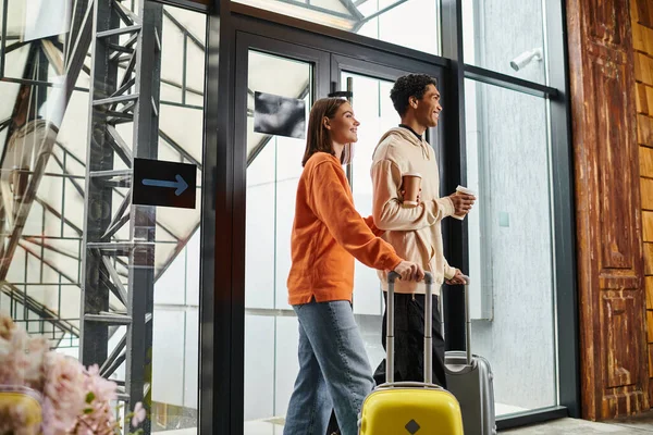 Happy diverse couple with travel luggage smiling and entering a modern hostel, holding coffee to go — Stock Photo