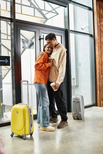 Amare coppia diversificata con caffè che abbraccia accanto a valigie da viaggio in un ingresso moderno di ostello — Foto stock