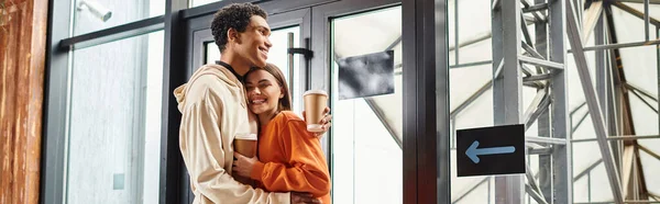 Happy diverse travel couple with coffee embracing beside modern glass entrance of hostel, banner — Stock Photo