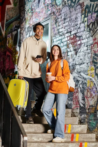 A cheerful diverse couple standing on stairs with colorful graffiti, one holding a yellow suitcase — Stock Photo