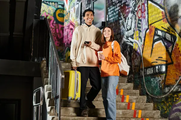 Heureux couple diversifié debout sur les escaliers avec des graffitis sur le mur, homme noir tenant une valise jaune — Photo de stock