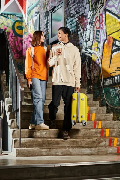 Joyful diverse couple standing on stairs with graffiti wall, black man holding a yellow suitcase — Stock Photo