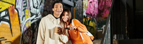 Happy couple in casual wear with coffee cup and smartphone near graffiti painted wall, banner — Stock Photo