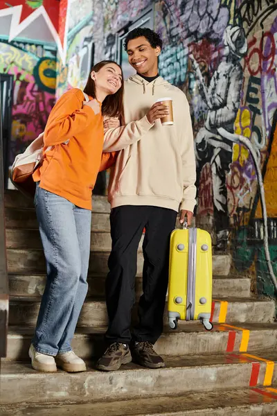 Різноманітна пара посміхається і стоїть поруч один з одним на сходах з графіті, кавою і багажем — стокове фото