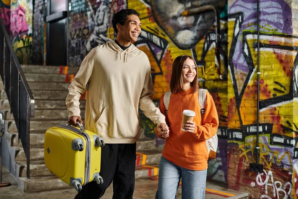 Cheerful couple holding hands and walking with a yellow suitcase in a graffiti-painted wall, hostel — Stock Photo