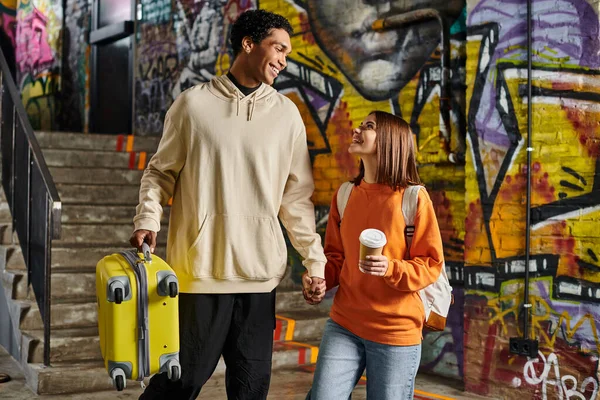 Diverse couple holding hands and smiling with a yellow suitcase in a graffiti-painted wall, hostel — Stock Photo