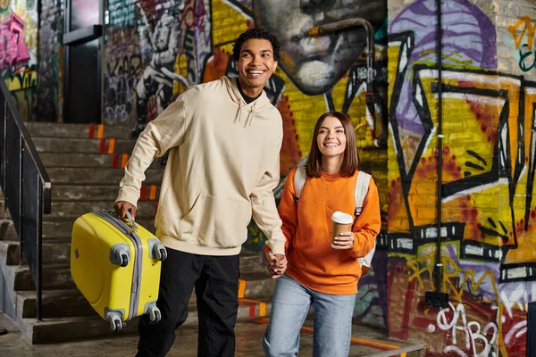 Diverse couple holding hands and smiling with a yellow luggage in a graffiti-painted wall, hostel — Stock Photo