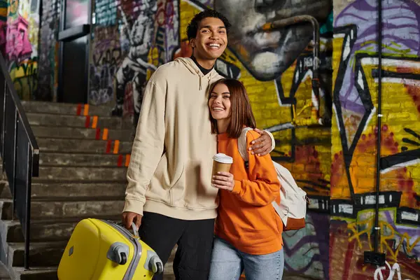 Feliz pareja diversa abrazando por una escalera con graffiti en el fondo, hombre negro con equipaje - foto de stock