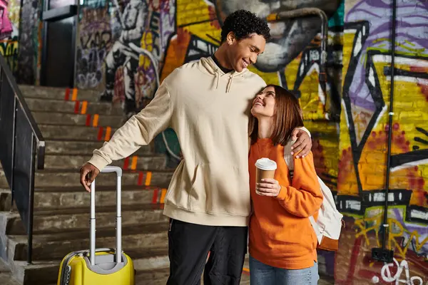 Feliz casal diversificado abraçando por parede com graffiti no fundo, homem preto com bagagem — Fotografia de Stock
