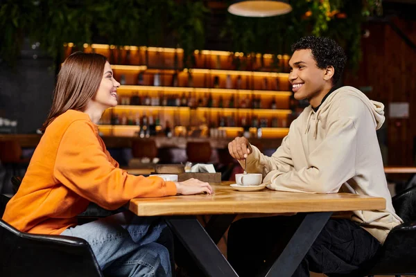 Веселая женщина и черный мужчина, сидящие в уютном кафе, вступая в дружеский разговор за чашками кофе — стоковое фото