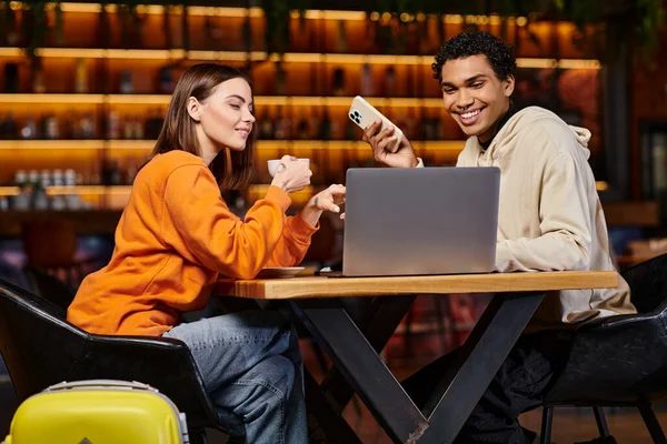 Feliz casal diverso olhando para laptop em um café moderno, mulher segurando xícara de café perto de homem preto — Fotografia de Stock