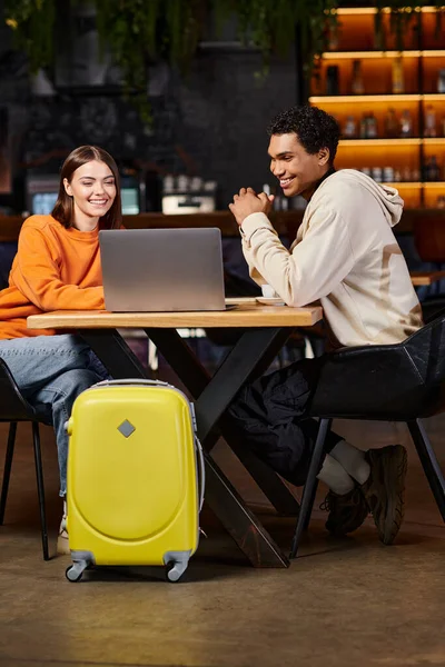 Feliz pareja diversa mirando a la computadora portátil, hombre negro y mujer sentado en la mesa cerca de equipaje de viaje - foto de stock