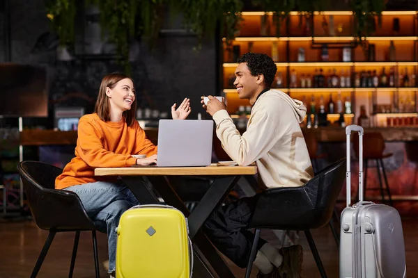 Homme et femme noirs riant et parlant à une table de café, avec leurs valises à côté d'eux, ordinateur portable — Photo de stock