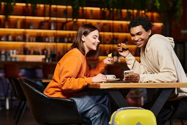 Diversa pareja almorzando y compras en línea desde el teléfono inteligente su equipaje en la cafetería albergue - foto de stock