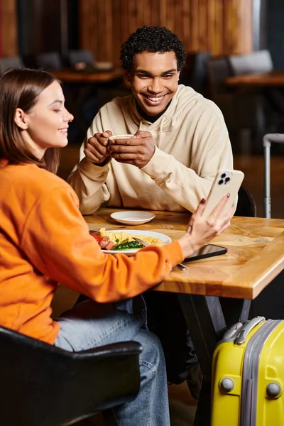 Uma mulher sorridente vestida com roupas coloridas mostrando smartphone para namorado preto com xícara de café — Fotografia de Stock