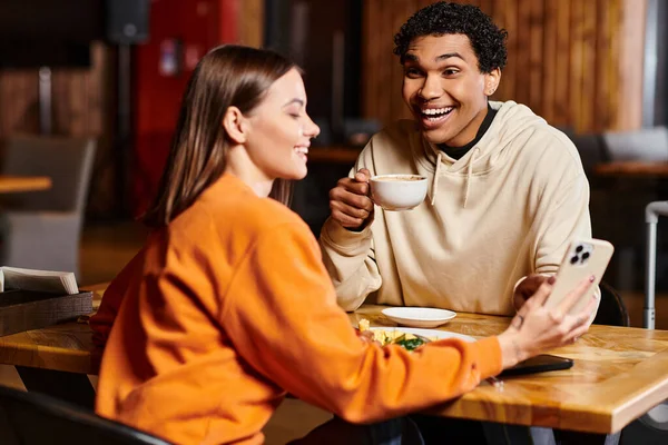 Веселий чорний чоловік, дивлячись на жінку, сидить за дерев'яним столом зі смартфоном у місцевому кафе, час обіду — стокове фото