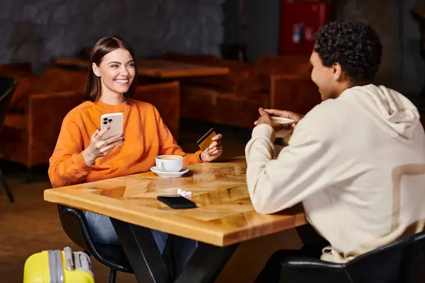 Diverse couple sharing quiet conversation at table, woman holding smartphone and credit card — Stock Photo