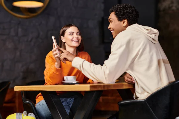 Mujer feliz en suéter naranja muestra el teléfono al novio negro sonriente mientras está sentado en la cafetería - foto de stock