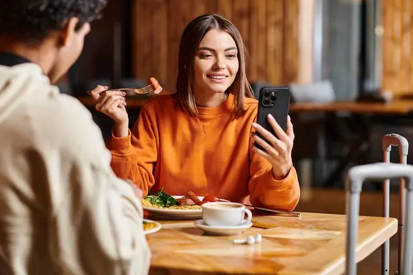 Young woman in vibrant orange sweater happily looking at her phone near black boyfriend during meal — Stock Photo